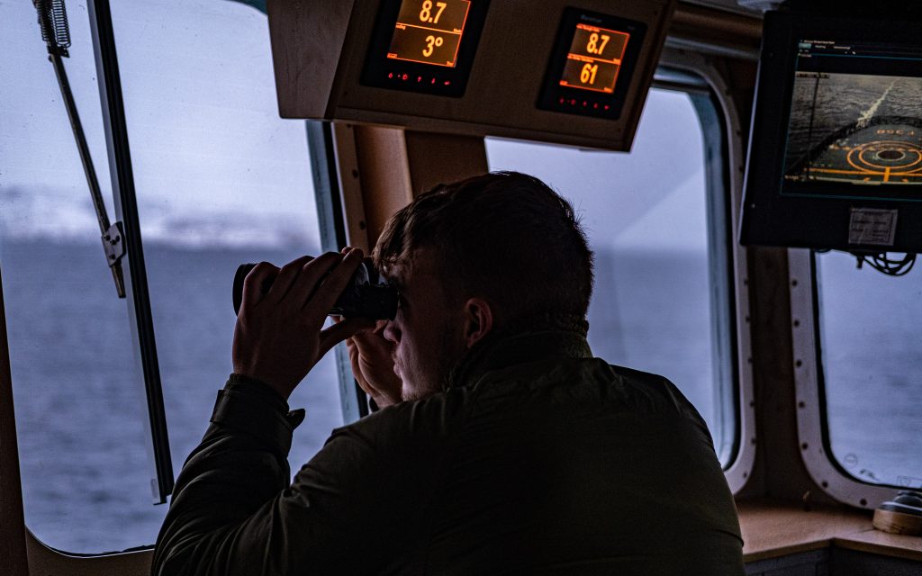 Photo: A Royal Danish Navy crew member on board the HDMS Triton looks through their binoculars. The Royal Danish Navy frigate HDMS Triton is part of the Joint Arctic Command Denmark, a joint operational territorial command of the Danish Defence operating in Greenland and the Faroes Islands. The command’s main task is surveillance and military defence of Greenland and the Faroe Islands. It is also responsible for fisheries control, search-and-rescue, environmental monitoring and logistical support to civil society. Their capabilities provide a vital contribution to the security and defence of the High North. Credit: NATO via Flickr. https://flic.kr/p/2ojszag