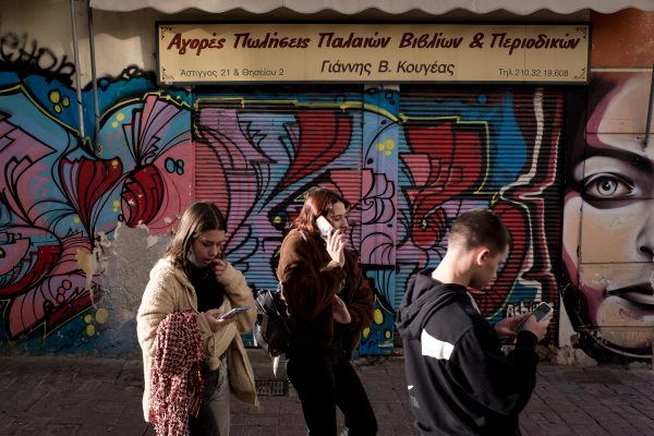 Photo: Three European people are holding their phones looking at an app while they are passing front of a tech graffiti in the center of Athens, Greece on December 8, 2021. Credit: Nikolas Kokovlis/NurPhoto.