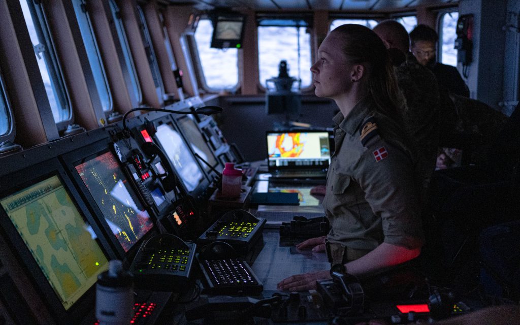 Photo: A Royal Danish Navy crew member inspects their surroundings in the bridge of the HDMS Triton as it patrols around Greenland.
The Royal Danish Navy frigate HDMS Triton is part of the Joint Arctic Command Denmark, a joint operational territorial command of the Danish Defence operating in Greenland and the Faroes Islands. The command’s main task is surveillance and military defence of Greenland and the Faroe Islands. It is also responsible for fisheries control, search-and-rescue, environmental monitoring and logistical support to civil society. Their capabilities provide a vital contribution to the security and defence of the High North. Credit: NATO via Flickr https://flic.kr/p/2ojrd6X.